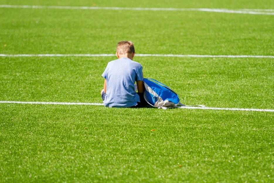 A child sits alone on a bright green turf field next to a blue backpack. This evocative image is placed near text about abuse within athletics.
